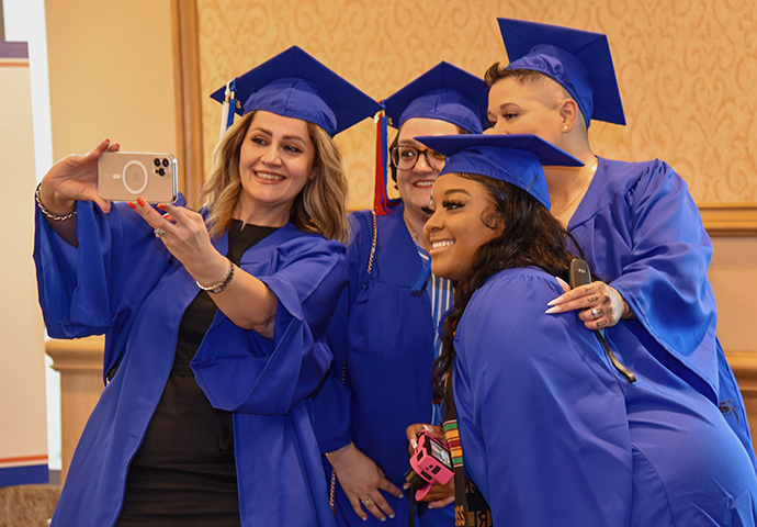 Four graduates in cap and gown pose for a selfie together.