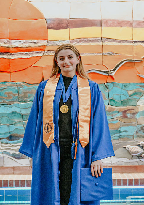 Person stands in cap and gown in front of sunset mural